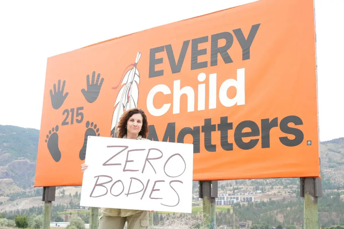 Dallas Brodie holding a 'Zero bodies' placard in front of an Every Child Matters billboard on Penticton Indian Band land, October 2025.